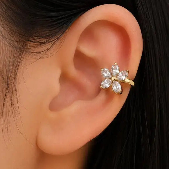 A close-up of a person's ear wearing a gold ear cuff in the shape of a flower with clear rhinestones.
