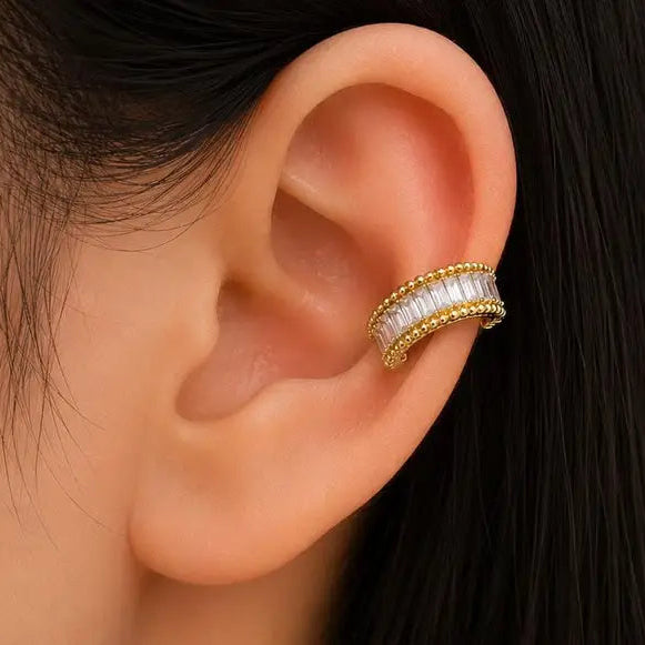 A close-up of a person's ear wearing a gold ear cuff adorned with a row of clear baguette gemstones.