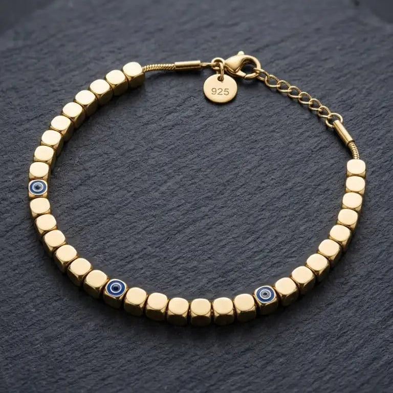 A gold beaded bracelet with small evil eye charms, displayed on a dark slate surface.