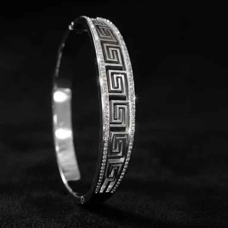A silver-toned hinged bangle bracelet with a Greek key pattern and pavé-set clear stones, photographed on a dark textured background.