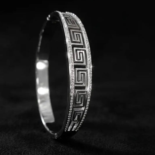 A silver-toned hinged bangle bracelet with a Greek key pattern and pavé-set clear stones, photographed on a dark textured background.
