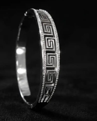 A silver-toned hinged bangle bracelet with a Greek key pattern and pavé-set clear stones, photographed on a dark textured background.