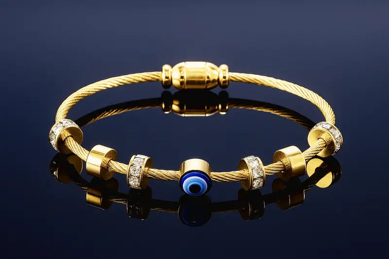 A gold cable-wrapped bracelet with gold spacer beads, some adorned with clear crystals, and a central blue evil eye charm, displayed on a reflective dark surface.