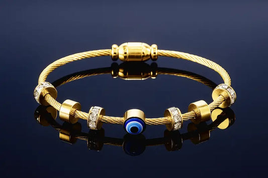 A gold cable-wrapped bracelet with gold spacer beads, some adorned with clear crystals, and a central blue evil eye charm, displayed on a reflective dark surface.